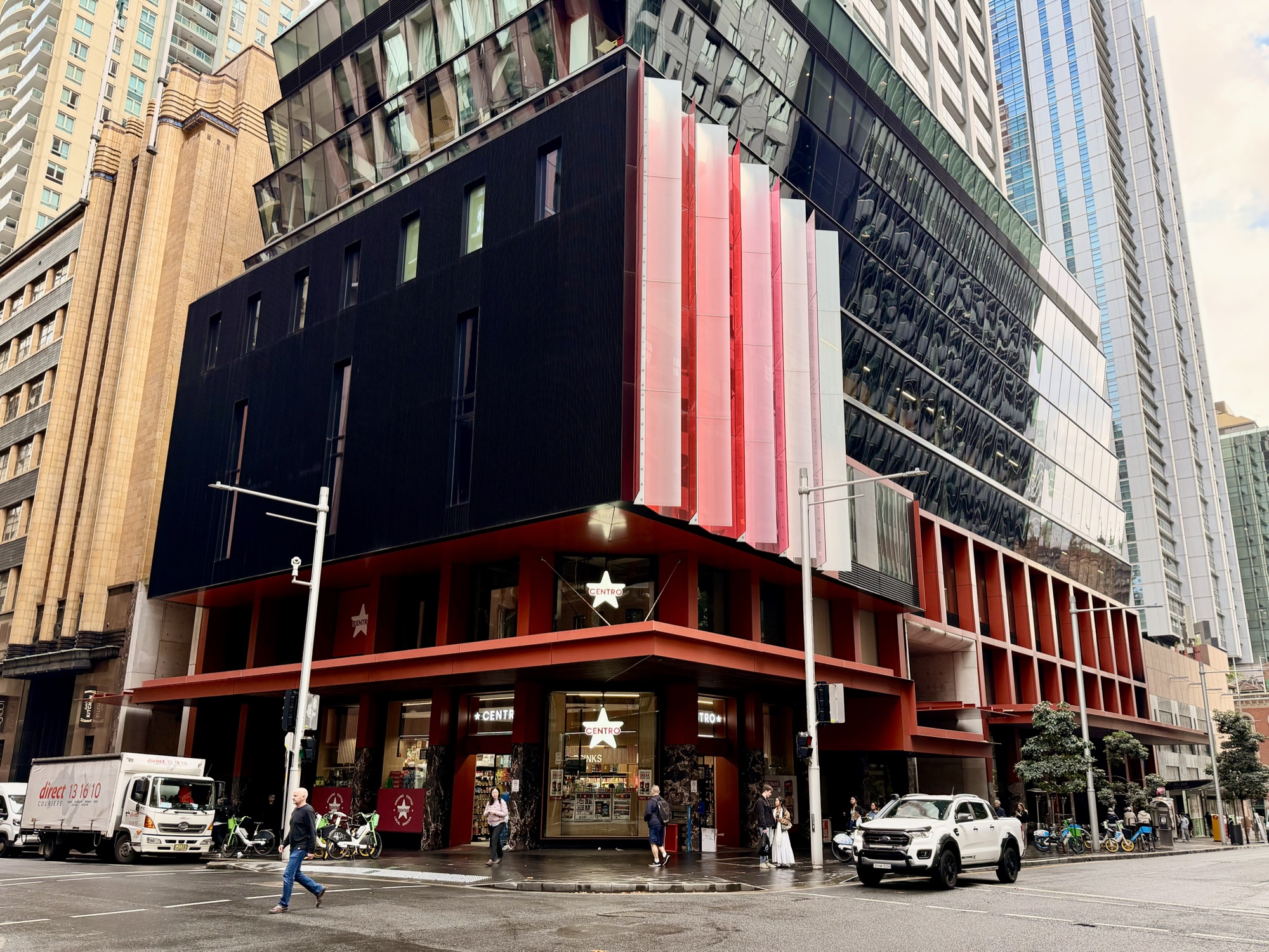 A tall dark building with a red-striped corner facade, on a busy city street with taller towers behind