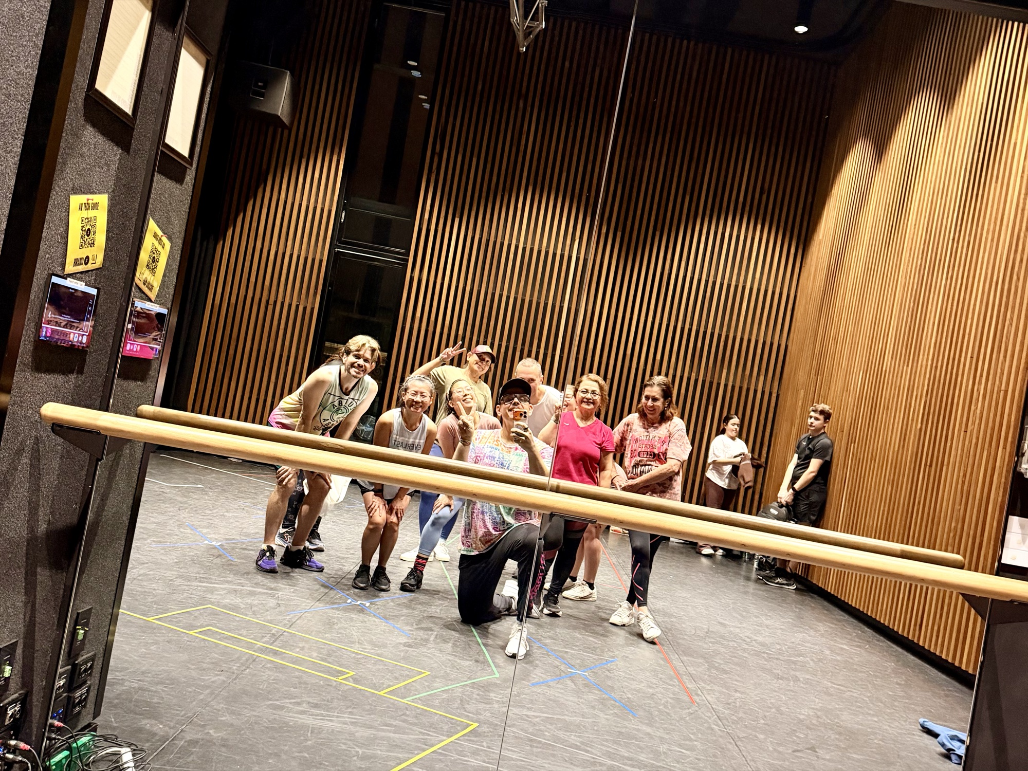 A group of dancers strikes a playful pose together, reflected in a large studio mirror with a ballet barre across the front