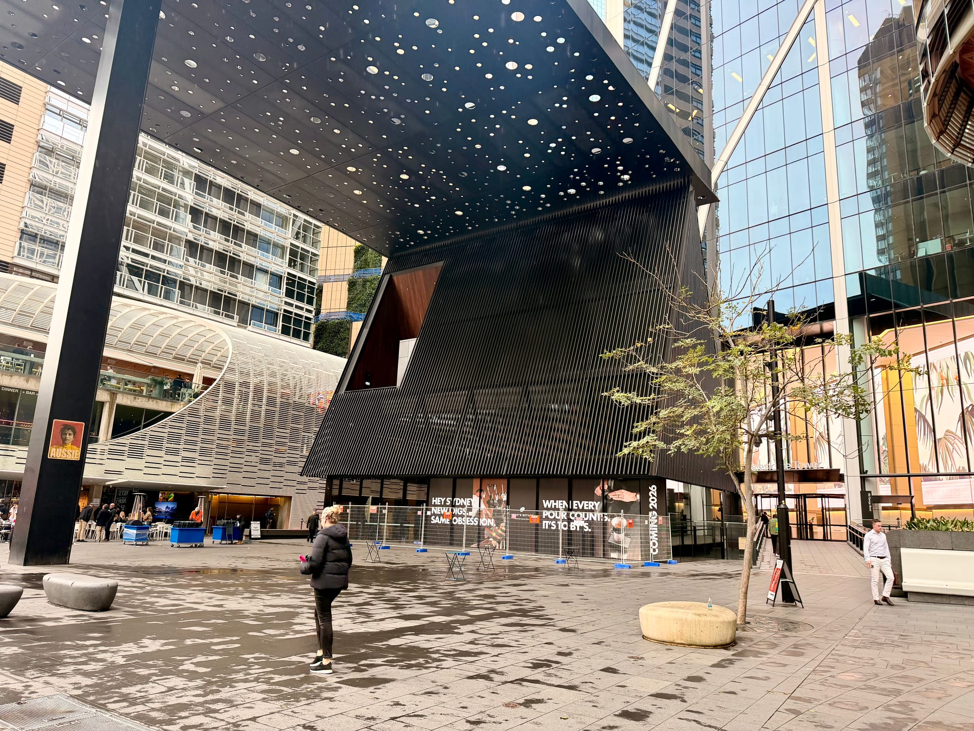 A striking black building with a faceted, perforated facade rising over an open city plaza, glass towers behind