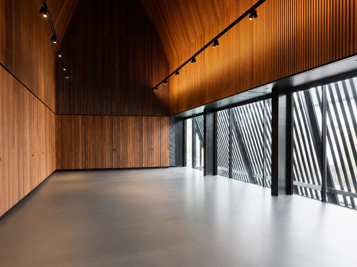 A bright, empty room with warm wood-panelled walls, a high angled timber ceiling and a polished concrete floor, with full-height glass along one side