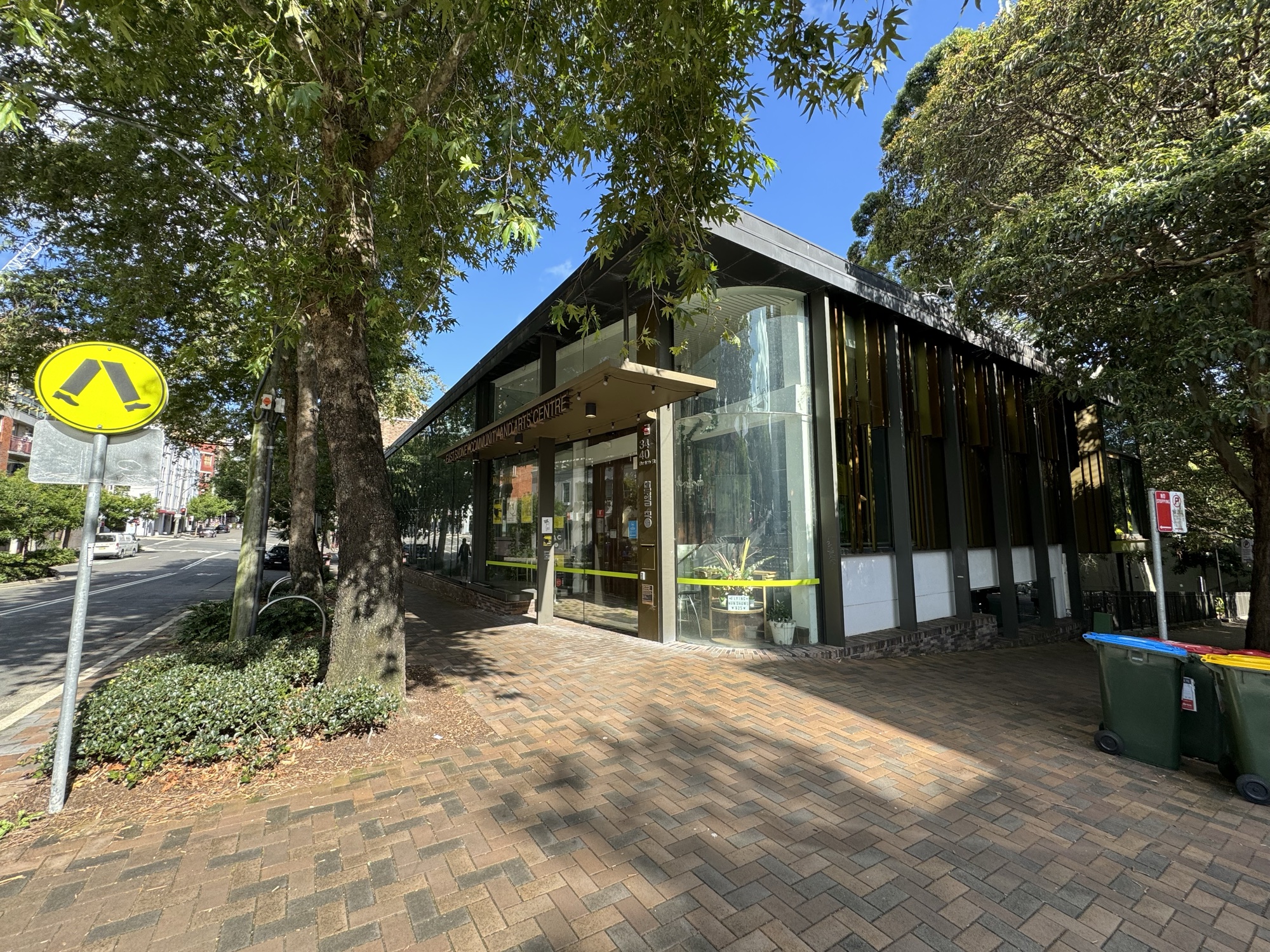 A modern building with a steep angled roof and tall glass walls, beside a big tree on a quiet corner