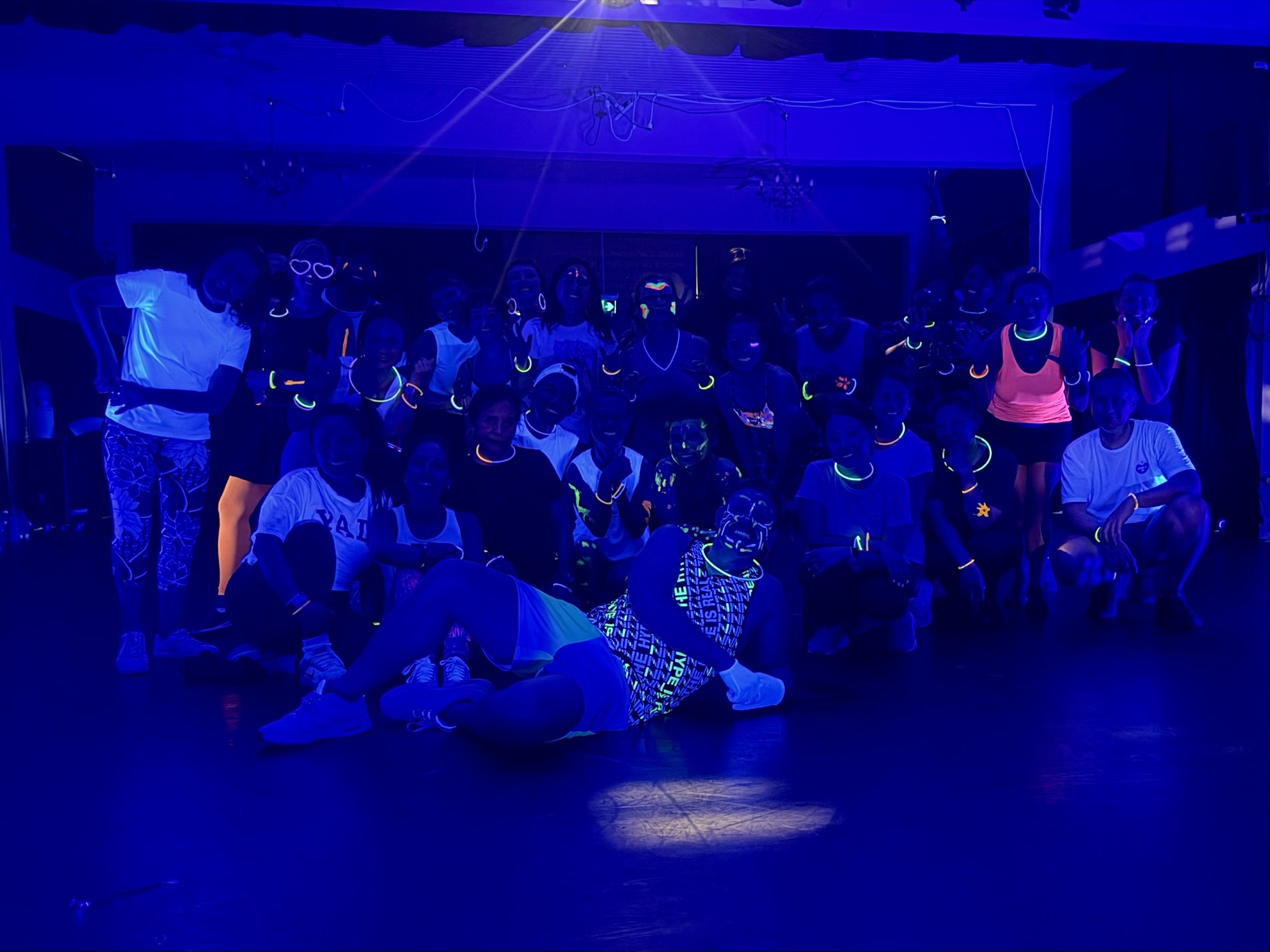 A big group glows in deep blue UV light, wearing bright glow-stick necklaces and streaks of neon face paint